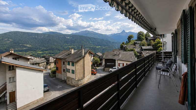 Balcony with a view of a village and mountains, featuring a table and chairs, and an awning.