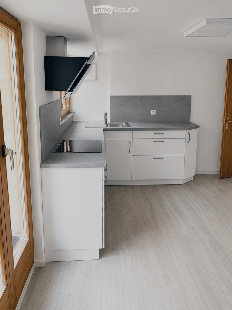 Modern kitchen with white cabinets, grey countertops, and a stainless steel sink. Features a range hood and wooden flooring.