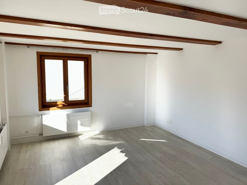 Empty room with light wooden flooring, white walls, and a large window with wooden frame. Exposed wooden beams on the ceiling.