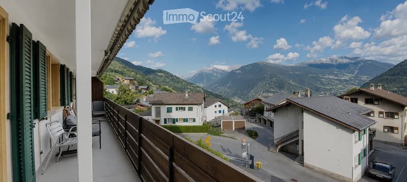 Balcony with wooden railing, overlooking a scenic mountain valley with houses and a road below.