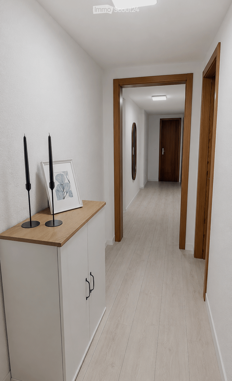 Modern hallway with light wooden flooring, white walls, and a small wooden console table with a framed picture and two tall candles. The hallway leads to another room with a visible wooden door and a round mirror on the wall.