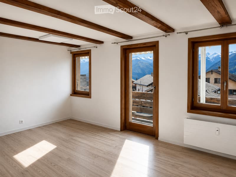 Empty room with wooden flooring, white walls, exposed wooden beams on the ceiling, large windows and a sliding door leading to a balcony with mountain views.