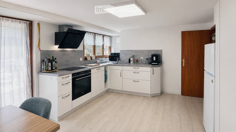 Modern kitchen with white cabinets, stainless steel appliances, and a wooden floor. Features include a refrigerator, oven, sink, coffee maker, and a small dining area with a chair and table.