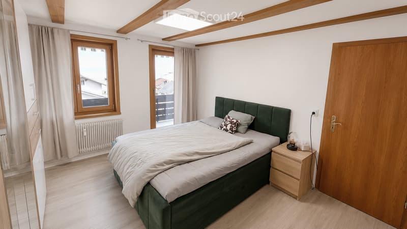 Cozy bedroom with wooden floors, large window, green upholstered headboard, nightstand with lamp, and radiator.
