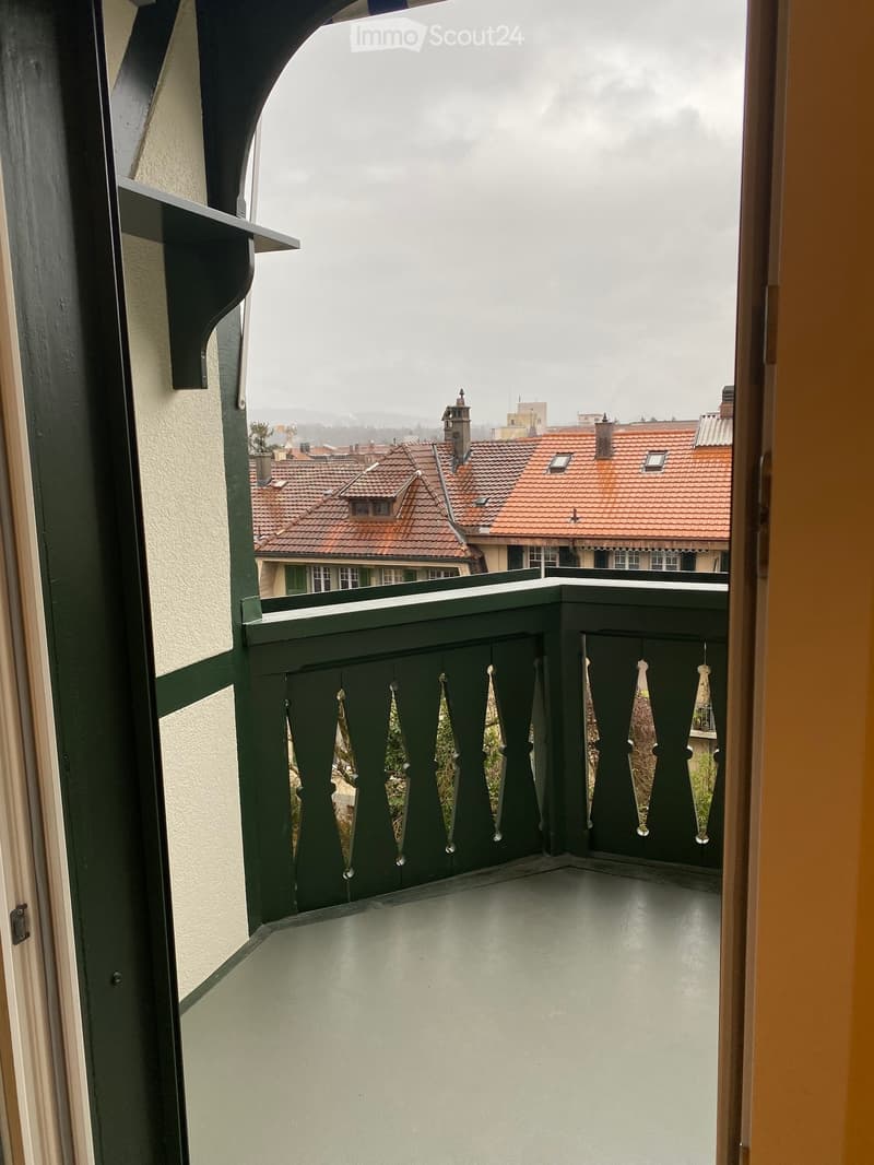 Balcony with green metal railing and decorative balusters, overlooking a cityscape with red-tiled roofs and chimneys.