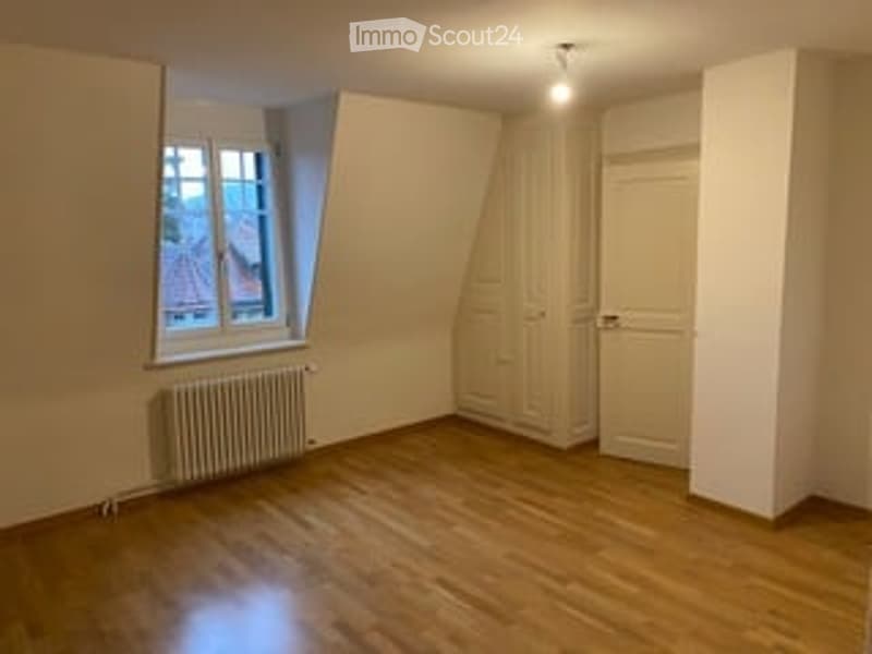 Completely empty room with wooden flooring, white walls, a window with a view of a roof, a radiator, and a closed door.