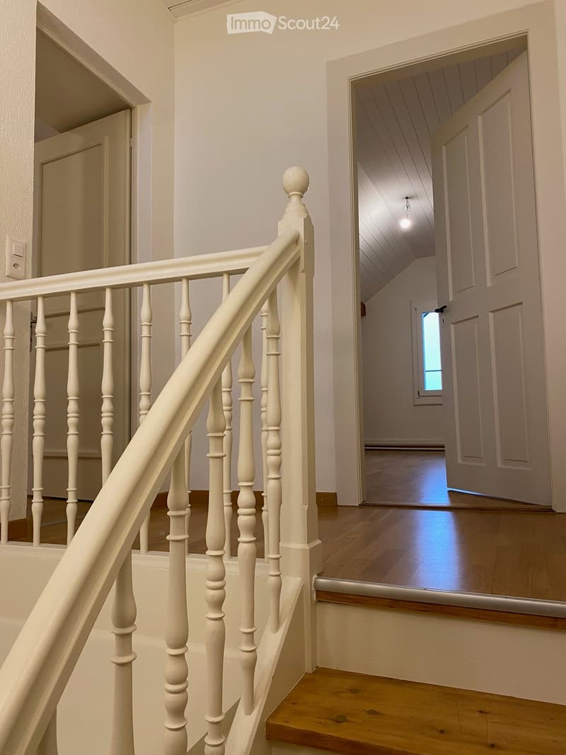 Wooden staircase with white balustrade and handrail, leading to a hallway with wooden flooring and white walls.