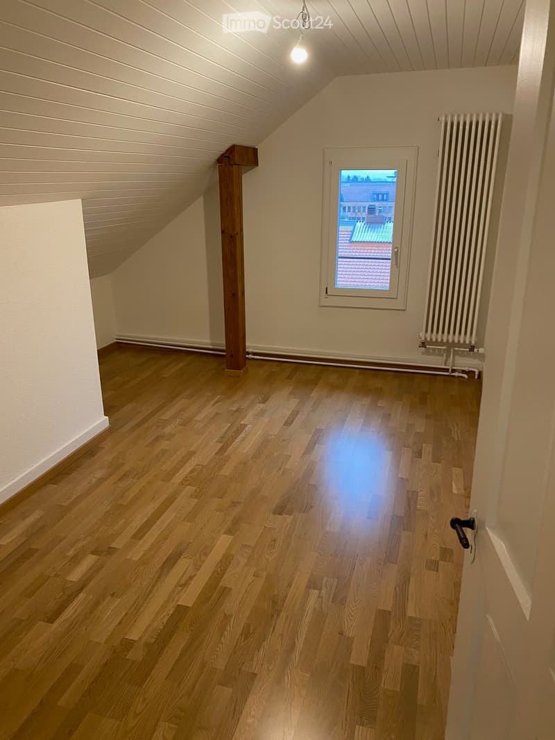 Empty room with wooden flooring, white walls, and a window showing a view of a roof and building outside. A radiator is visible near the window.