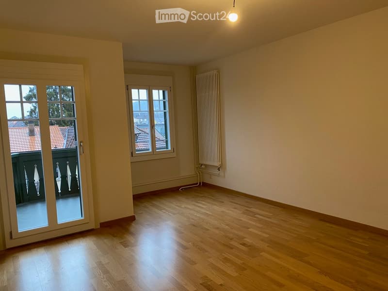 Empty room with wooden flooring, large windows, and a radiator on one wall. The room has a sliding glass door leading to a balcony.
