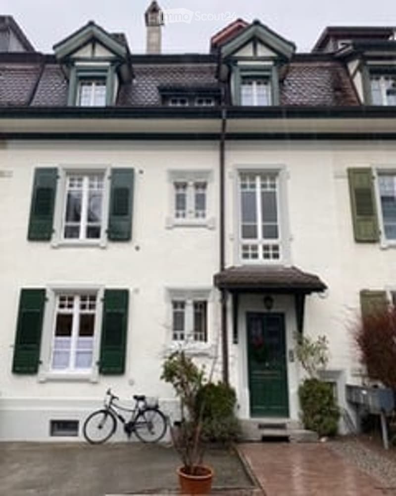 A classic European-style building with white walls, green shutters, and multiple windows. A bicycle is parked near the entrance, and a small potted plant is placed beside the door.