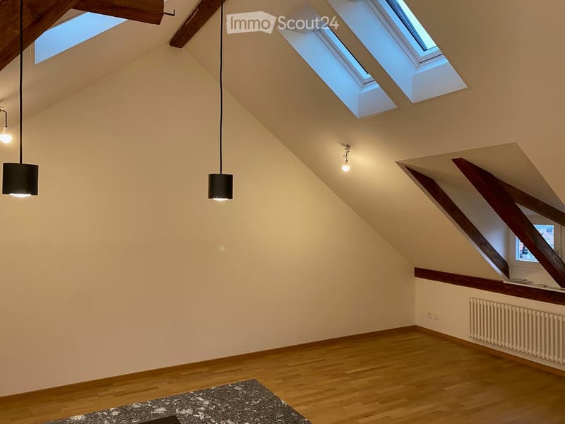An attic room with wooden floors, exposed wooden beams, skylights, and pendant lights. The space is empty, with no furniture or furnishings.