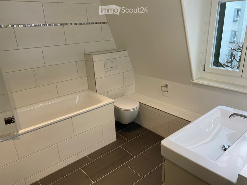 Modern bathroom with a bathtub, toilet, and sink, featuring light-colored tiles and a window with a view of a tree.
