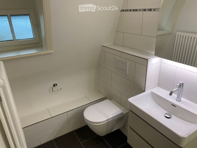 Modern bathroom with a toilet, sink, and cabinet. Features a radiator, mirror, and a window with frosted glass.