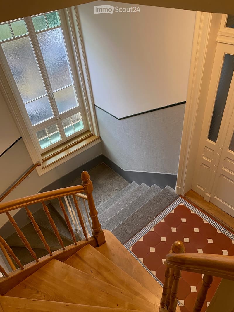 Wooden staircase with a decorative red and grey tiled landing, featuring a wooden handrail and balusters, with a large window on the left side.