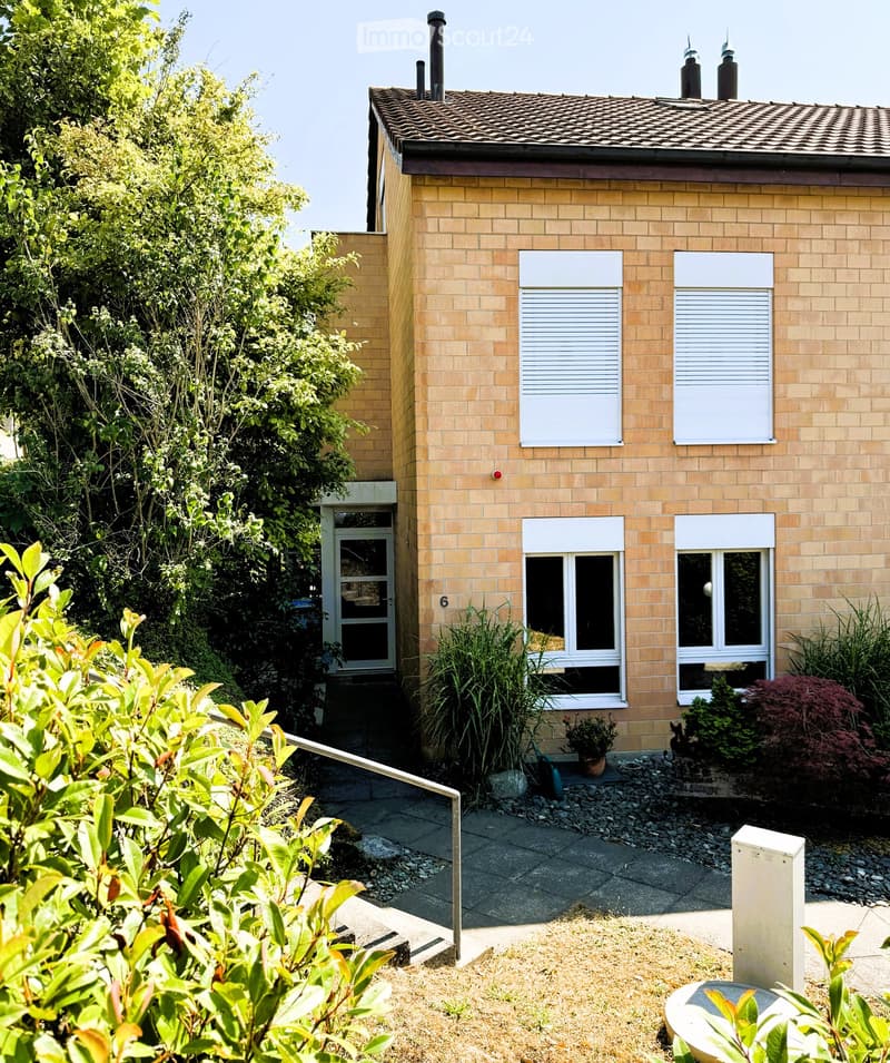 Two-story house with beige brick exterior, multiple windows with white frames, and a small garden area with plants and a concrete pathway leading to the entrance.