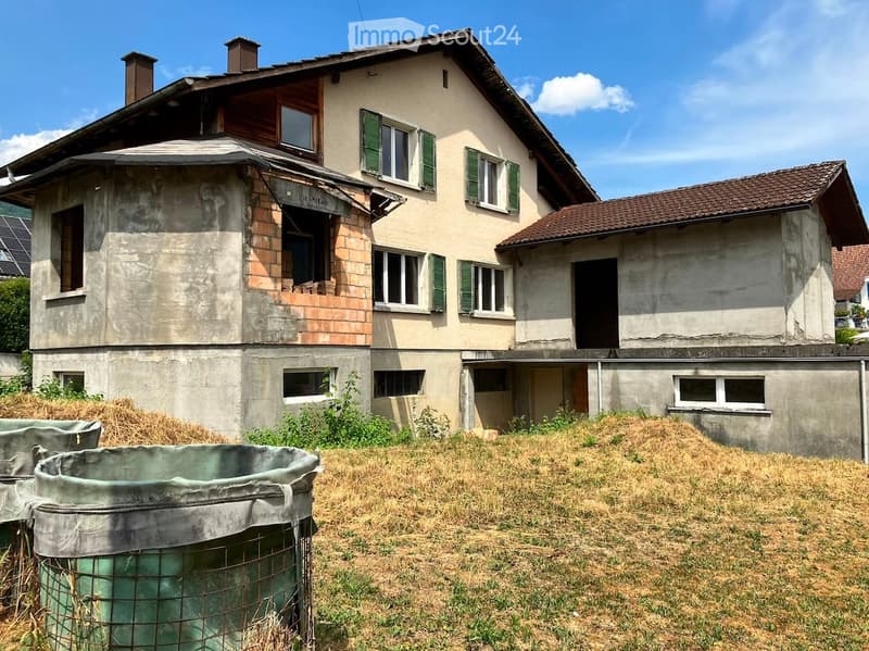 Two-story house with beige walls, green shutters, and a partially overgrown yard with hay bales and a large metal container.