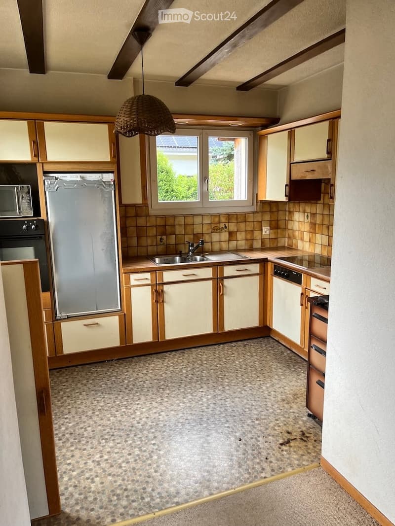 The kitchen features light-colored cabinets with wooden trim, a speckled tile floor, and a window above the sink. Appliances include a refrigerator, oven, microwave, and dishwasher. A hanging woven pendant light is visible.