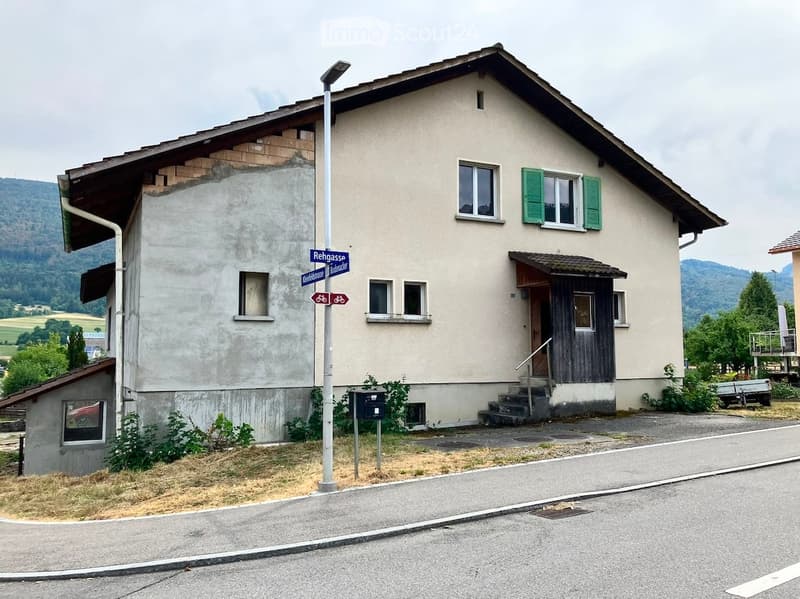 Two-story house with beige walls, green shutters, and a wooden entrance. The roof shows exposed wooden beams on one side. A street sign reading "Rehgasse" is visible in front.