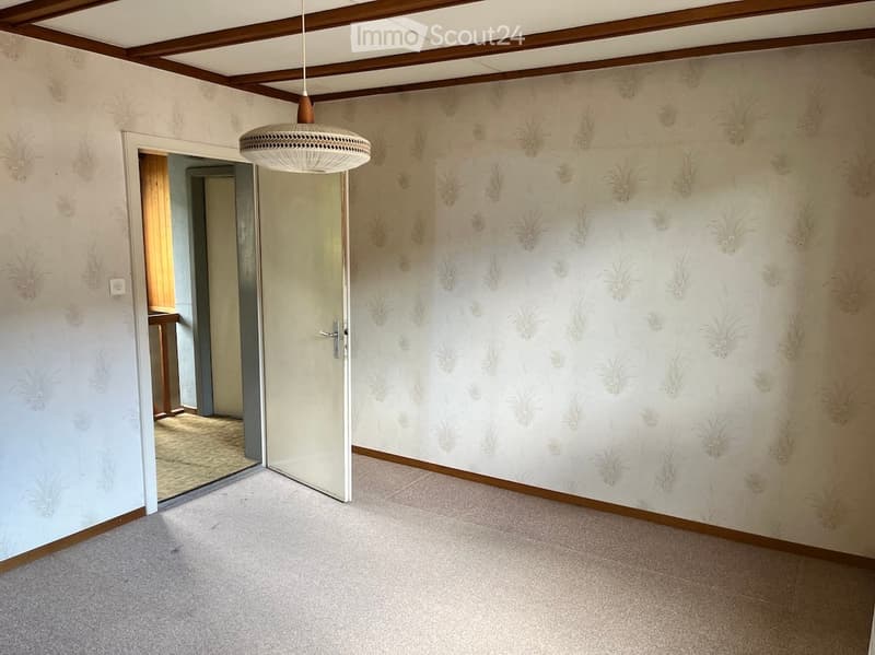 Empty room with light-colored carpet, patterned wallpaper, and a hanging light fixture. The room has a wooden door leading to another space.