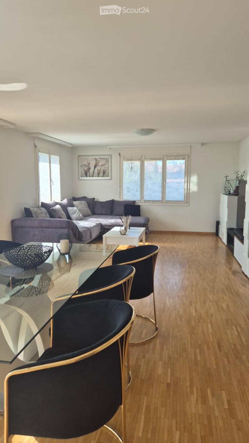 Modern living room with light-colored walls, wooden flooring, large windows, grey sectional sofa, white coffee table, and black chairs with gold accents.