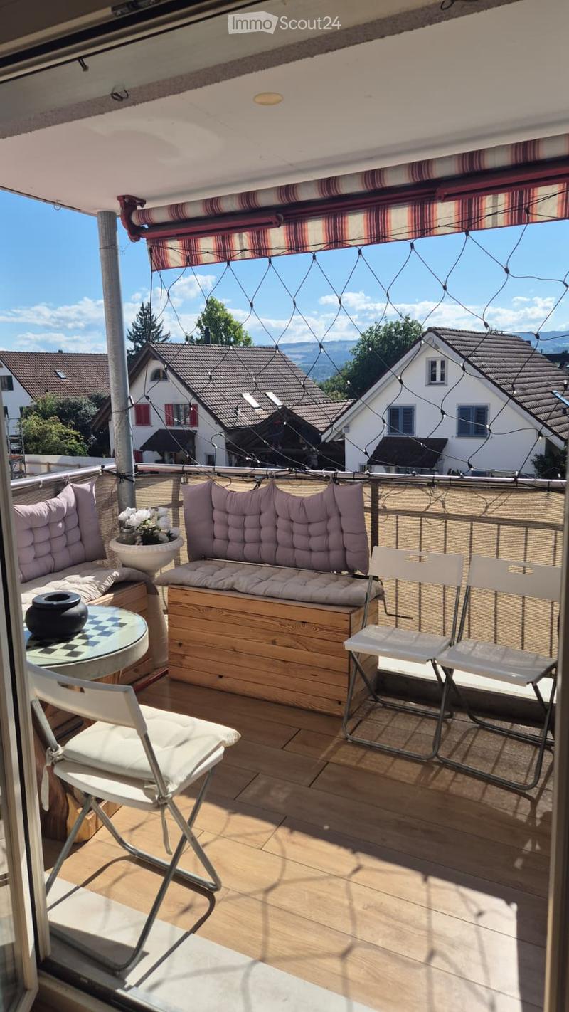Balcony with wooden floor, cushioned seating, round table, and chairs, overlooking a residential area with houses and mountains.