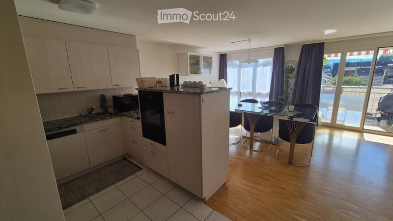 Open-plan kitchen and dining area with modern cabinets, a central island, and a dining table with chairs. Large windows and a sliding glass door lead to a balcony.