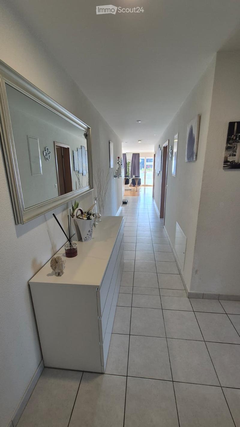 Clean hallway with tiled flooring, white walls, and a small console table with decorative items. Mirrors and framed artwork are mounted on the walls, leading to an open living area with furniture visible at the end.