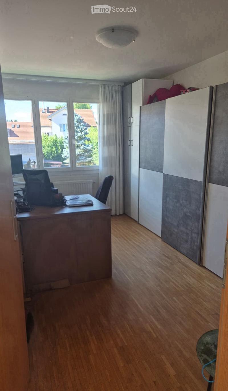 A furnished room with wooden flooring, a large window with curtains, a desk with a laptop and chair, and a refrigerator with items on top.