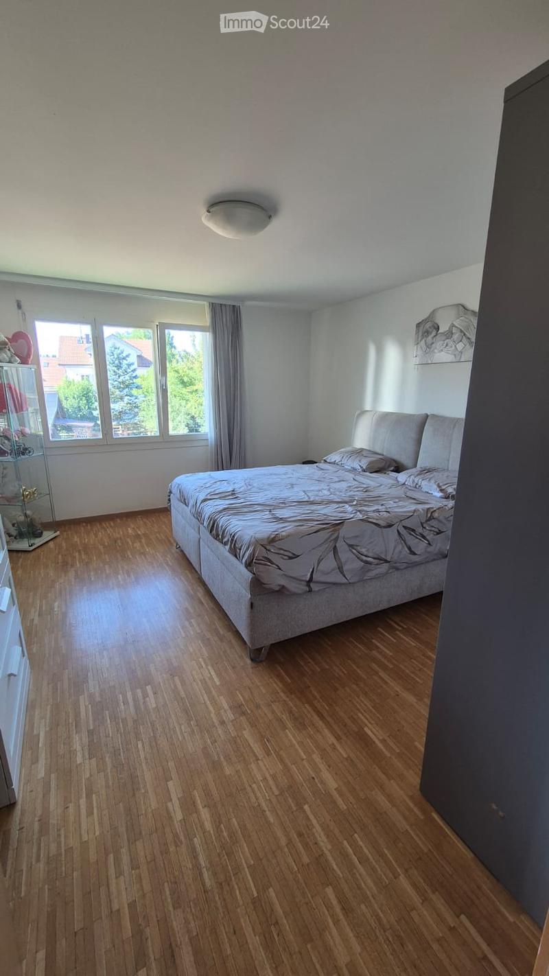 Bright bedroom with wooden flooring, large windows with curtains, a neatly made bed, and a nightstand with a lamp.