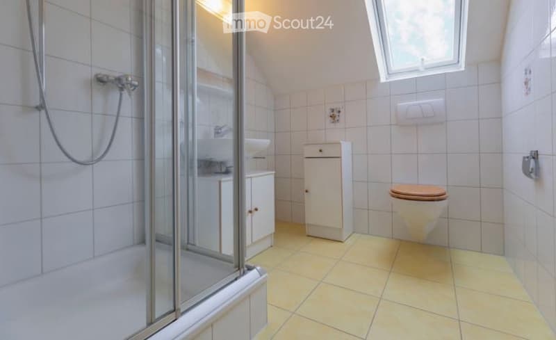Bright bathroom with white tiled walls and yellow tiled floor, featuring a toilet, sink, and a glass-enclosed shower area.