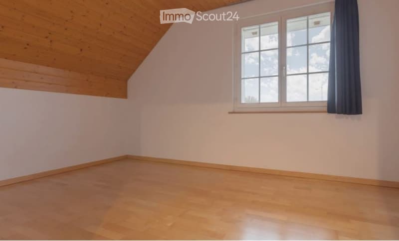 Empty room with wooden flooring, white walls, and a large window with dark curtains. The ceiling is slanted with wooden paneling.