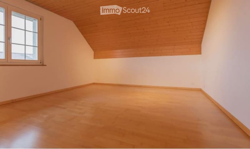 Empty room with wooden flooring and white walls, featuring a slanted wooden ceiling and a window on one side.