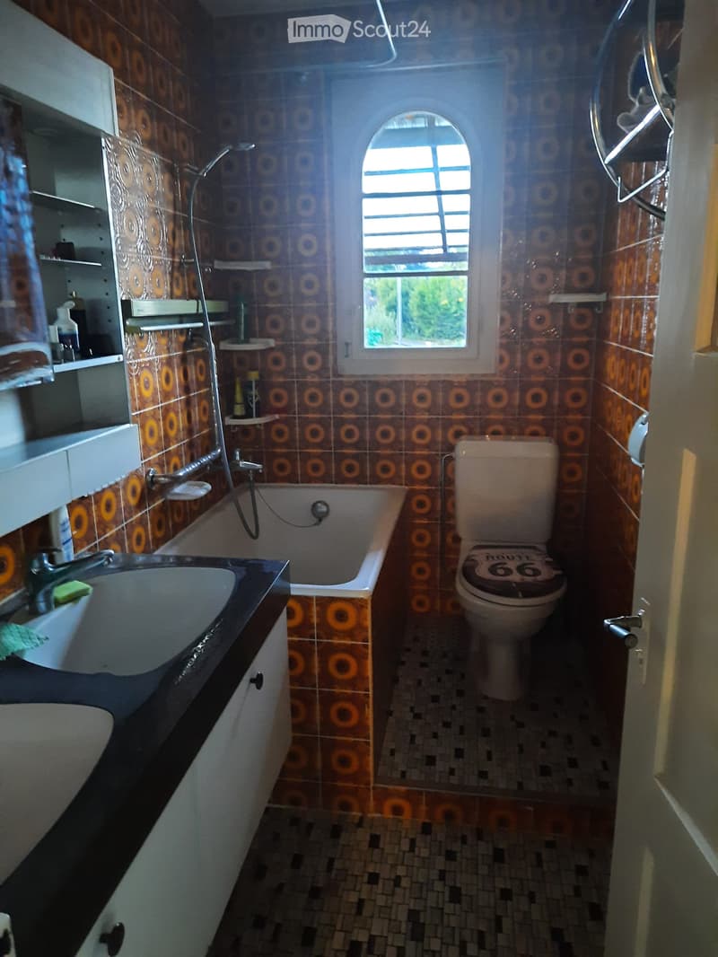 Bathroom with a sink, bathtub, toilet, and shelves. The walls are tiled with a circular pattern, and there is a window above the bathtub.