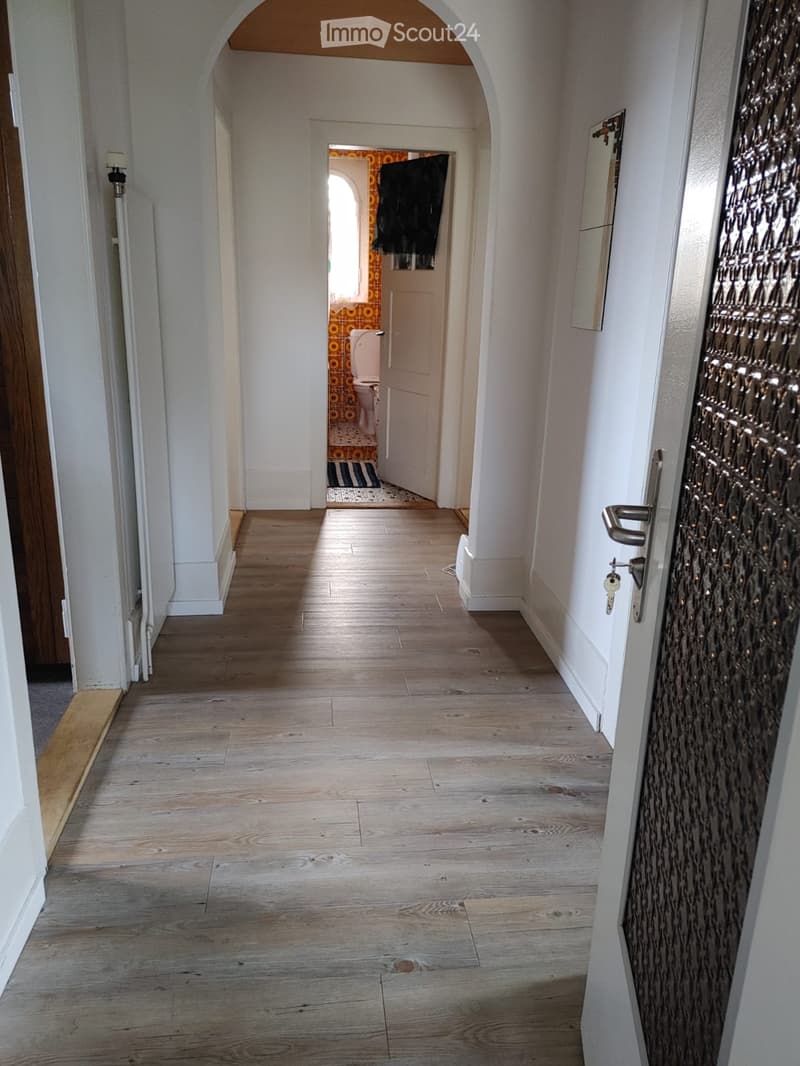 A long, narrow hallway with light wooden flooring, white walls, and an arched ceiling. A door on the right leads to a room with visible tiles and a toilet, while a mirror and a decorative door are visible on the right side.