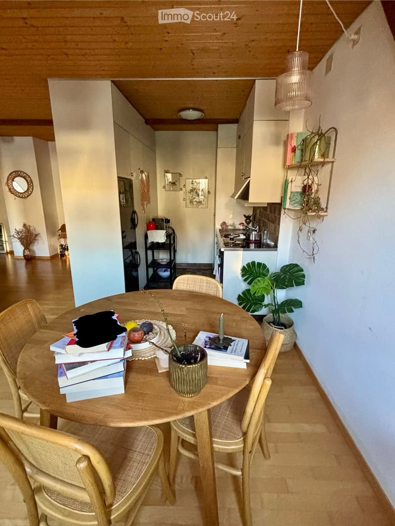 A cozy, open-plan living and dining area with a round wooden table surrounded by four wicker chairs. The table holds books, a vase with a plant, and a candle. A kitchen area is visible in the background with a stove, cabinets, and a small shelf with items