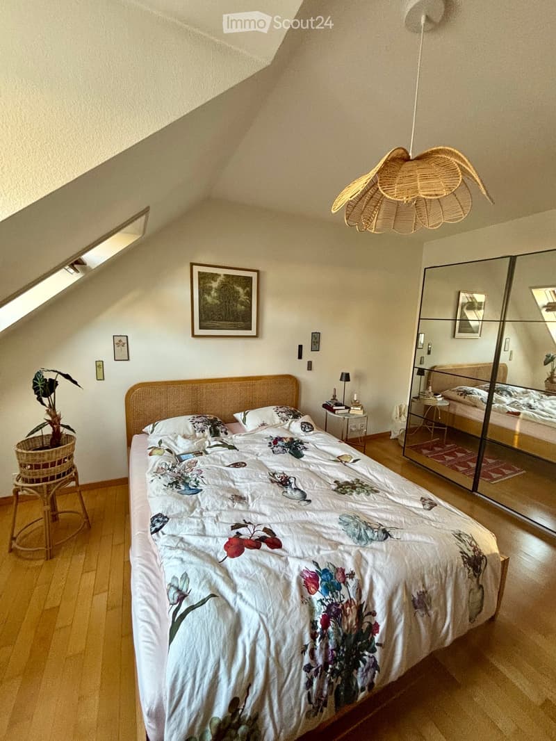 Cozy attic bedroom with wooden floors, a large bed with floral bedding, a wicker headboard, a nightstand with a lamp, a mirror, and a small plant on a wicker stool.
