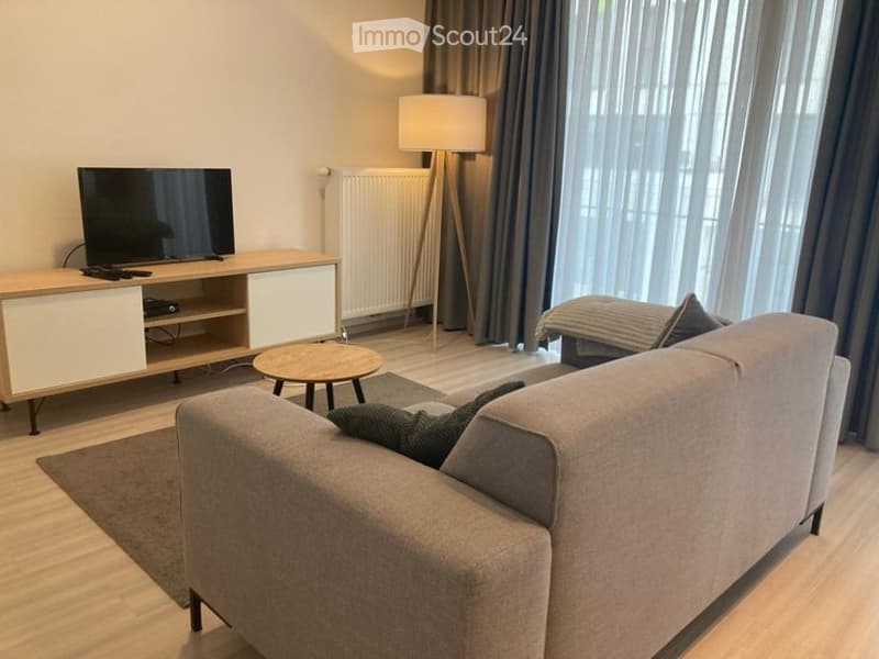 Modern living room with a grey sofa, coffee table, TV stand, floor lamp, and ottoman. Large windows with grey curtains and a radiator are visible.