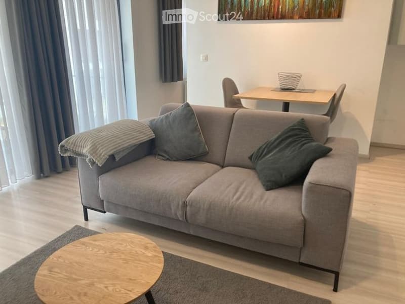 A modern living room with a grey sofa, wooden coffee table, dining table, and chairs. The room features large windows with sheer curtains and a cozy, minimalist design.