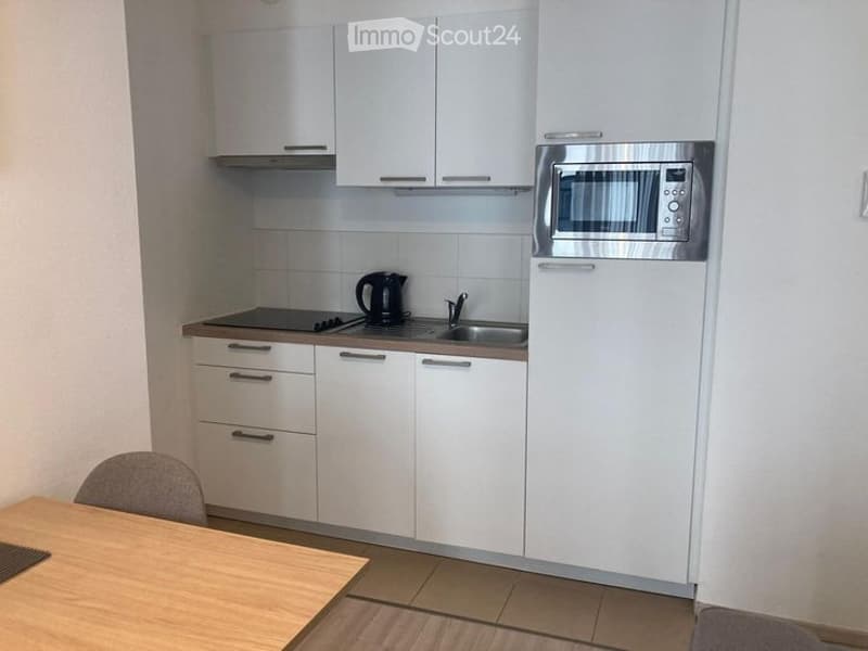 Modern kitchen with white cabinets, stainless steel microwave, refrigerator, stove, sink, and a small wooden dining table with a chair.