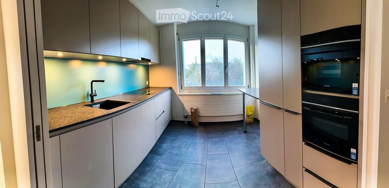Modern kitchen with white cabinets, granite countertop, stainless steel sink and faucet, and built-in ovens. Large windows provide natural light.