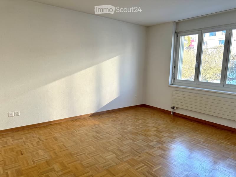 Empty room with wooden parquet flooring, white walls, and a large window with a radiator below it.