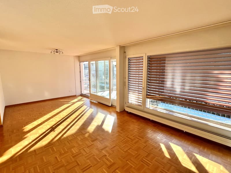 Empty room with wooden flooring, large windows with blinds, and a sliding glass door leading to a balcony.