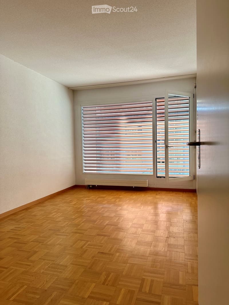 Empty room with wooden parquet flooring, light-colored walls, and a large window with blinds. Radiator visible below the window.