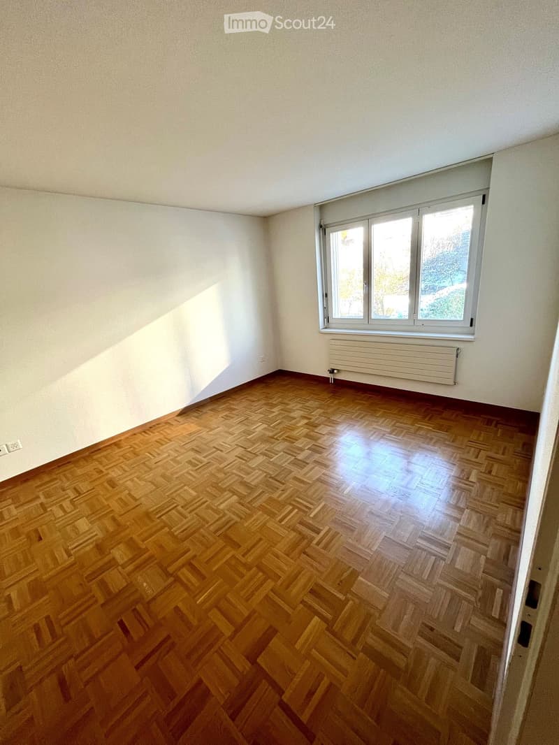 Empty room with wooden herringbone flooring, white walls, and a large window with a radiator below it.