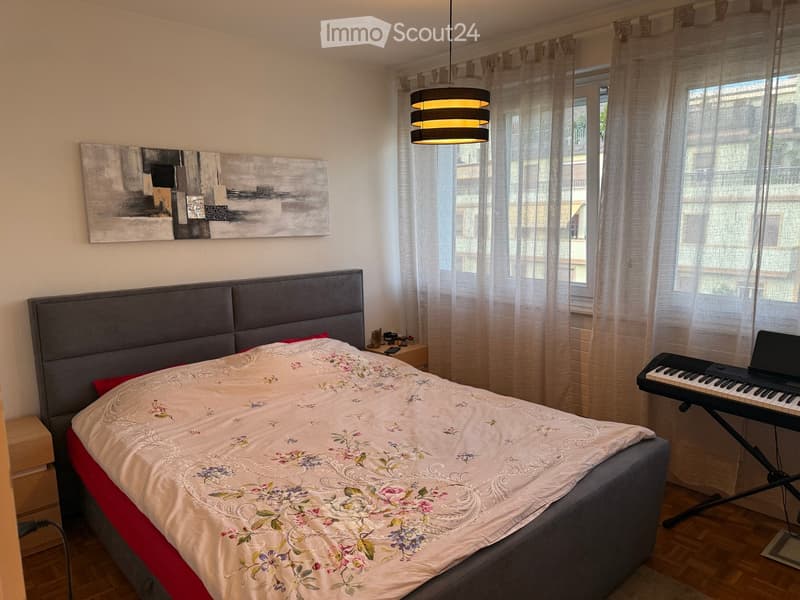 A well-lit bedroom with a large bed, a keyboard and laptop on a stand, a nightstand with small items, and a modern painting on the wall.