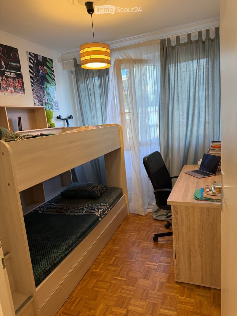 Cozy bedroom with a wooden bunk bed, a desk with a laptop, a chair, and a lamp hanging from the ceiling. The room features wooden flooring and large windows with curtains.