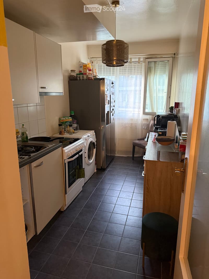 The kitchen features dark tiled flooring, white cabinets, and various appliances including a refrigerator, oven, washing machine, and stove. There is a small table with a chair, a hanging light fixture, and a window with curtains.