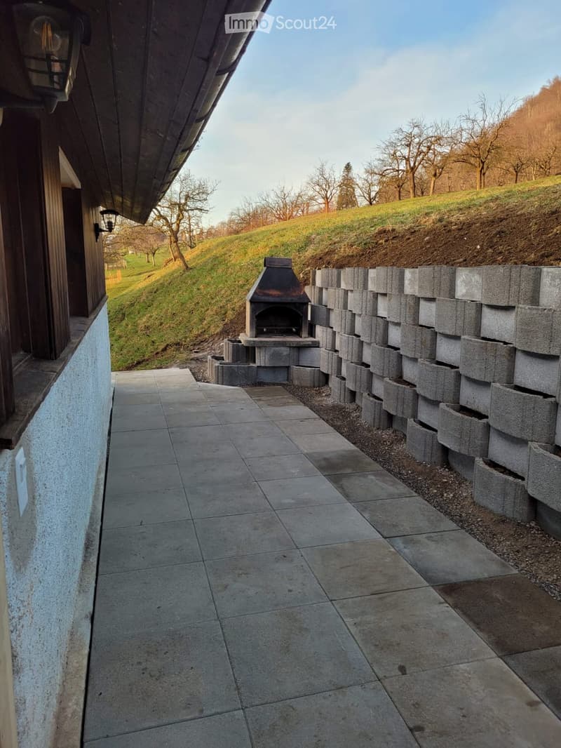 Outdoor patio area with tiled flooring, stone barbecue, and a stone wall, overlooking a grassy hill with trees.