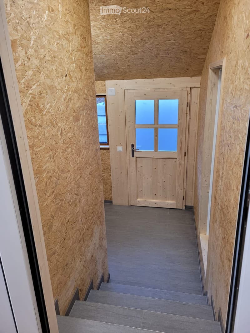 A narrow hallway with wooden walls, grey tiled floor, and a wooden double door with glass panels at the end. A small window is visible on the left side.