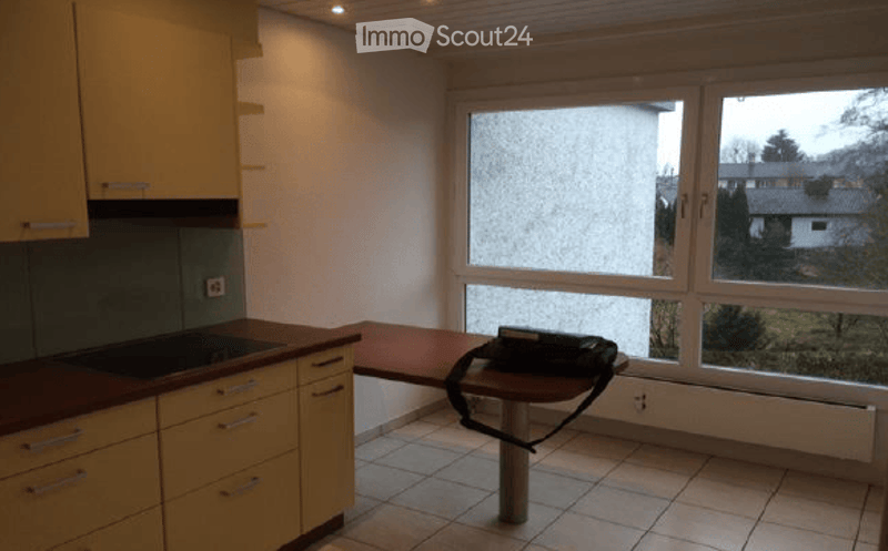A small, unfurnished kitchen with light-colored cabinets, a sink, and a small table with a chair. The room has tiled flooring and large windows offering a view of the outside.